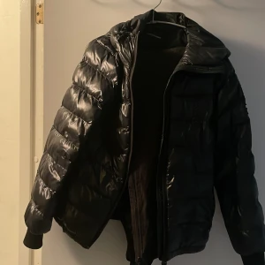Svart dunjacka från Peak Performance - Säljer en svart dunjacka från Peak Performance med huva och dragkedja. Jackan har en quiltad design och logotyp på ärmen. Perfekt för kyliga dagar och håller dig varm med sin vaddering. Säljer bort för den är för kort för mig