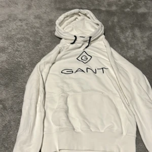 Vit hoodie från Gant - Säljer en vit hoodie från Gant med svart logotyp och text på bröstet. Tröjan har huva med dragsko och en stor magficka framtill. Perfekt för en avslappnad stil. Funkar till båda kön!