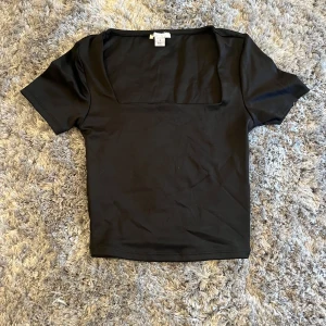 Svart topp med fyrkantig ringning - Svart lite kortare topp som är i ett lite glansigt snyggt tyg! Helt ny aldrig använd