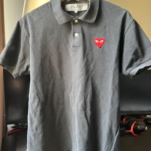 Svart pikétröja från Comme des Garçons Play - Säljer en klassisk svart pikétröja från Comme des Garçons Play med det ikoniska röda hjärtat broderat på bröstet. Tröjan har korta ärmar, krage och knappar framtill. Perfekt för en stilren och avslappnad look.