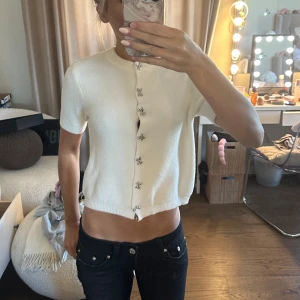 Vit kortärmad stickad topp med dekorativa knappar - Säljer en söt vit stickad topp med korta ärmar och glittriga dekorationsknappar framtill. Modellen är croppad och har en rund halsringning. Perfekt att styla till jeans eller kjol.