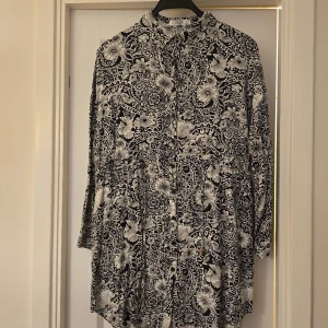 Svartvit blommig klänning från Lusa - Säljer en svartvit klänning från Lusa med blommigt och paisley-inspirerat mönster. Klänningen har lång ärm, knappar framtill och ett knytband i midjan för en snygg siluett. Perfekt för dig som gillar mönstrade plagg med lite extra detalj.