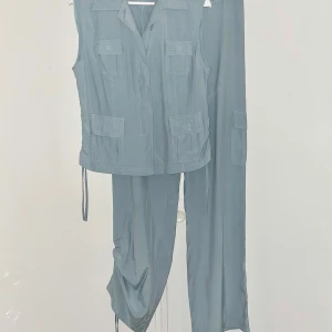 Ärmlös skjorta och byxor - Set bestående av en ärmlös skjorta och ett par byxor i matchande färg. Skjortan har en klassisk krage, knappstängning framtill och fickor. Byxorna har en avslappnad passform med dragsko nedtill för justerbar längd. Bystvidd 102cm. Midja/höft 82cm