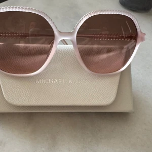Rosa solglasögon från Michael Kors - Snygga solglasögon från Michael Kors med stora, rundade bågar i ljusrosa plast och matchande rosa tonade glas. Perfekt accessoar för att lyfta din stil.