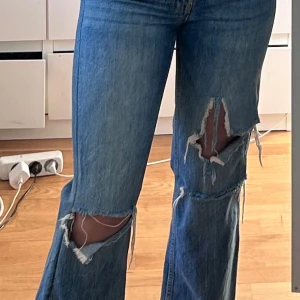 Blå slitna jeans från Gina Tricot - Säljer ett par blå jeans från Gina Tricot med raka ben och stora slitningar på båda benen. Jeansen har klassisk femficksdesign, normal passform och är tillverkade i denim. Perfekta för en avslappnad och trendig look.