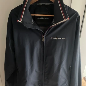 Marinblå vindjacka från Sail Racing - Säljer en marinblå vindjacka från Sail Racing med dragkedja och broderade detaljer. Jackan har hög krage med röd och vit rand, samt logga på bröstet och stor brodyr på ryggen. Perfekt för dig som gillar sportig stil. Sönder där man hänger jackan.