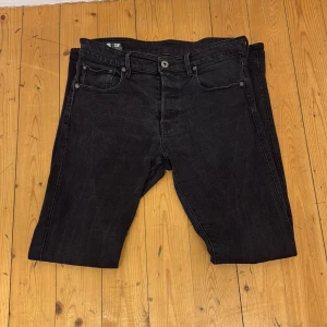 Svarta jeansbyxor G-Star RAW 3301 Straight - Svarta jeans från G-Star RAW, modell 3301 Straight. Klassisk femficksdesign med raka ben och normal passform. Jeansen har en diskret logotyp på bakfickan och läderpatch bak i midjan. Perfekta för en stilren och avslappnad look.