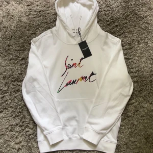 Vit hoodie från Saint Laurent - Snygg vit hoodie från Saint Laurent med färgglad broderad logga på bröstet. Klassisk huva med dragsko och magficka framtill. Perfekt för dig som vill ha en stilren men ändå unik look.