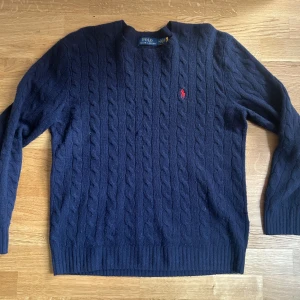 Mörkblå kabelstickad tröja från Polo Ralph Lauren - Kabelstickad mörkblå tröja från Polo Ralph Lauren med klassisk röd broderad logga på bröstet. Tröjan har rund halsringning och ribbade muddar vid ärmslut och nederkant. Perfekt för en stilren och tidlös look. Nypris:1700 mitt pris: 899. Köpt från Åhléns i Stockholm. Kom med frågor. Priset är ej hugget i sten  