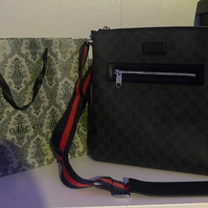 Messenger bag GG - Snygg svart axelväska från GG med diskret mönster och silverfärgad dragkedja framtill. Väskan har en bred axelrem i rött och blått, perfekt för att bära över axeln. Klassisk och stilren design med exklusiv känsla. Påsen på bilden ingår