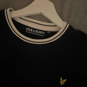 Mörkblå och vit t-shirt från Lyle & Scott - Snygg t-shirt från Lyle & Scott med mörkblå kropp och vita ärmar. T-shirten har rund hals med vit och blå randig kant samt den klassiska gula loggan broderad på bröstet.