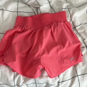 Rosa träningsshorts från Nike - Säljer ett par rosa träningsshorts från Nike med bred resår i midjan och klassisk logga på benet. Shortsen är tillverkade i ett lätt syntetmaterial och har en sportig passform som passar perfekt till träning.