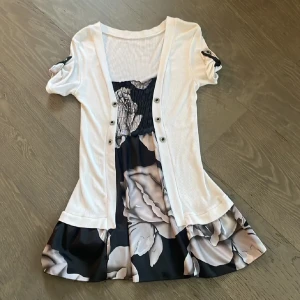 Kortklänning med blommigt mönster och vit kofta - Söt kortklänning med stora blommor i svart, vitt och grått. Klänningen har en fastsydd vit kofta med knappar framtill och korta ärmar med knappdetalj. Perfekt för dig som gillar en feminin och stilren look. Vintage miniklänning