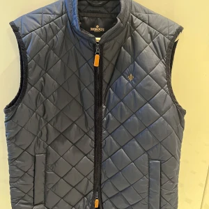 Mörkblå quiltad väst från Morris - Säljer en mörkblå quiltad väst från Morris med dragkedja framtill och diskret logga på bröstet. Västen har ståkrage och två fickor framtill. Perfekt för lager-på-lager-stil under kyligare dagar.