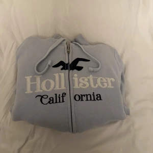 Ljusblå hoodie med dragkedja från Hollister - Mysig ljusblå hoodie från Hollister med dragkedja och broderad logga på bröstet. Tröjan har huva med dragsko och är fodrad med mjuk, vit teddy på insidan. Perfekt för kalla dagar.