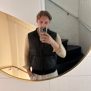 Svart dunväst från Polo Ralph Lauren - Säljer en svart dunväst från Polo Ralph Lauren med dragkedja framtill och diskret röd logga på bröstet. Västen har en klassisk passform och är perfekt för lager-på-lager-stil. Materialet är polyester med dunfyllning för extra värme. Sitter mer som S