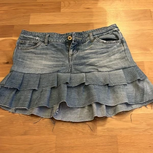 Jeanskjol med volanger - Säljer en  jeanskjol med dubbla volanger och snygga detaljer på fickorna. Kjolen har klassiska fickor fram och bak samt knapp och dragkedja i midjan. Perfekt för dig som gillar en cool och trendig stil.