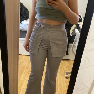 Grå kostymbyxor med unik detalj - Säljer ett par grå kostymbyxor med hög midja och speciell överlappande detalj framtill. Byxorna har raka ben och stängs med dragkedja och knapp. Perfekta för dig som vill sticka ut med en modern twist på klassiska byxor.