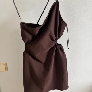 Brun one shoulder klänning Jazz cutout - Säljer en brun one shoulder klänning i modellen Jazz cutout. Klänningen har smal axelrem och asymmetrisk design med lätt rynk vid sidan. Perfekt för dig som gillar stilrena och trendiga plagg.