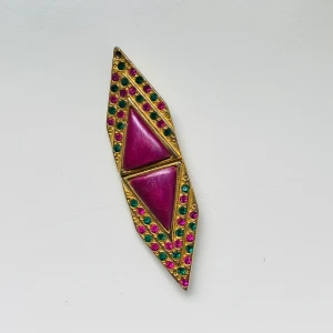 Vintage brosch i guld med färgglada stenar - Unik brosch i guldtonad metall med två stora triangulära lila stenar i mitten. Omgiven av små runda rosa och gröna stenar som ger ett färgglatt och retro intryck. En sten saknas Broschen har en avlång, spetsig form och passar perfekt som statement-accessoar.