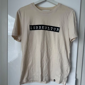 Odz merch Zubbkultur - Snygg beige t-shirt från Dunder Kläder med svartvit texttryck 'ZUBBKULTUR' på bröstet. Klassisk passform och rund halsringning. Perfekt för en avslappnad stil.