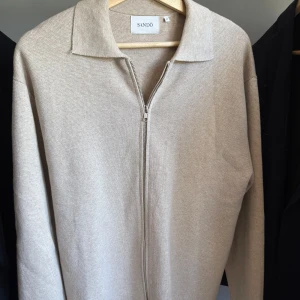 Beige stickad kofta med dragkedja från Sandö - Snygg beige stickad kofta från Sandö i storlek M. Tröjan har dragkedja framtill och klassisk krage. Aldrig använd men lappen är borttagen 