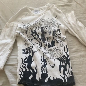 Vit långärmad tröja med anime-tryck - Säljer en vit långärmad tröja med svart anime-inspirerat tryck framtill. Motivet föreställer en tecknad tjej med stora ögon, långa tofsar och kjol, omgiven av eldliknande mönster och japansk text. Perfekt för dig som gillar unik streetstyle! 