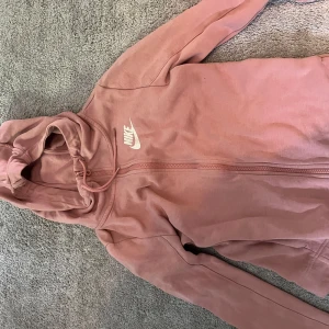 Rosa hoodiejacka från Nike - Säljer en rosa hoodiejacka från Nike med dragkedja framtill och huva med dragsko. Jackan har en enkel design med Nike-logga på bröstet och är perfekt för en avslappnad stil.