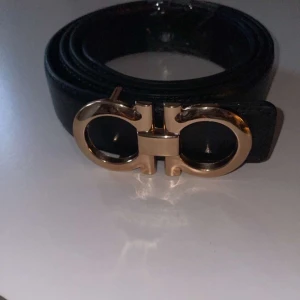 Svart bälte med guldfärgat spänne från Salvatore Ferragamo - Snyggt svart bälte i skinn med ett ikoniskt guldfärgat dubbel-Gancini-spänne från Salvatore Ferragamo. Perfekt accessoar för att lyfta din outfit med en lyxig touch. Bältet har en klassisk och stilren design.