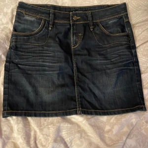 Mörkblå jeanskjol från b.young - Säljer en klassisk mörkblå jeanskjol från b.young med snygga kontrastsömmar och femficksmodell. Kjolen har bälteshällor och knapp- samt dragkedjestängning framtill. Perfekt till vardags med en t-shirt eller stickad tröja.