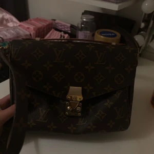 Brun axelväska  - Säljer en klassisk brun axelväska med  guldfärgat lås. Väskan har justerbar axelrem i läder och flera fack inuti för enkel organisering. Perfekt för dig som gillar lyxiga accessoarer.