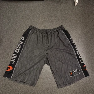 Svarta mesh shorts från GASP INC - Säljer ett par svarta mesh shorts från GASP INC med vita småprickiga detaljer och tryckta loggor längs sidorna. Shortsen har resår i midjan och är perfekta för gym eller träning.