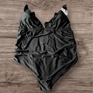 Svart baddräkt med volangdetaljer - Säljer en svart baddräkt från SWIM i storlek 3XL med volangdetaljer längs axelbanden och v-ringad hals. Baddräkten har justerbara axelband och är tillverkad i ett mjukt, stretchigt material som passar perfekt för bad.