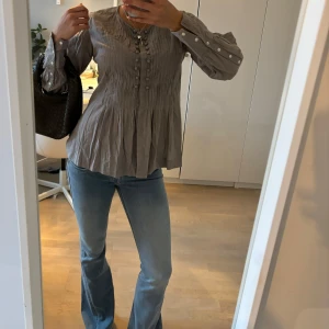 Grå blus från DAY Birger et Mikkelsen - Supersnygg grå blus från DAY Birger et Mikkelsen i storlek 36. Blusen har långa ärmar med knappar vid ärmslut, dekorativa knappar framtill och ett lätt rynkat tyg som ger en luftig känsla. Perfekt till jeans eller kjol.