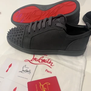 Gråa sneakers med nitar från Christian Louboutin - Säljer ett par svarta sneakers från Christian Louboutin med mattsvarta nitar på tån och den ikoniska röda sulan. Skorna har snörning och är tillverkade i mocka med diskreta sömmar och detaljer.