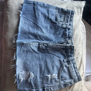 Mini Jeanskjol med slitningar och fransig kant  - Säljer en kort jeanskjol i klassisk blå denim med slitna detaljer och fransig nederkant. Kjolen har fickor både fram och bak samt bälteshällor. Perfekt för en avslappnad och trendig look.