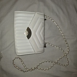 Vit quiltad axelväska med kedja - Säljer en stilren vit axelväska med quiltat mönster och guldfärgad kedja. Väskan har ett elegant spänne framtill och är perfekt för att lyfta din outfit. Rymmer det viktigaste och passar till många tillfällen.