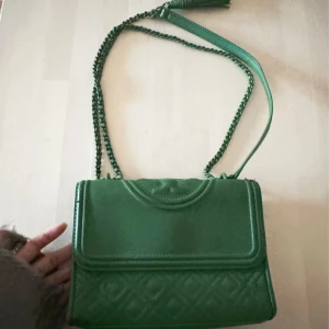Grön axelväska från Tory Burch - Snygg grön axelväska från Tory Burch med kedjerem och tofsdetalj. Väskan har quiltad yta och magnetlås, samt innerfack med dragkedja. Perfekt för dig som vill ha en färgklick till din outfit.