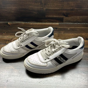 Vita sneakers från Polo med blå detaljer - Säljer ett par vita sneakers från Polo med blå ränder på sidorna och guldtext på hälen. Skorna har snörning, perforerad tå och platt sula. Perfekta för en sportig och avslappnad stil. 