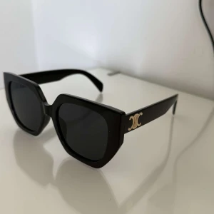 Svarta solglasögon Celine - Ett par Stilrena svarta solglasögon, en med markerad, fyrkantig båge och mörka glas. Sidorna pryds av den ikoniska guldloggan. 🌺