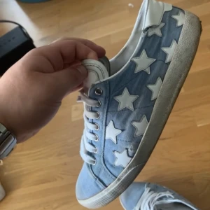 Saint Laurent sneakers med stjärnor - Säljer ett par blåa Saint Laurent sneakers med vita stjärnor på sidorna. Skorna har vit snörning, vit sula och är tillverkade i jeansmaterial. Perfekta för dig som vill sticka ut med en unik design.