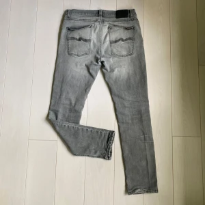 Grå jeans från Nudie Jeans - Säljer ett par grå jeans från Nudie Jeans med slitningar vid knäna och klassiska bakfickor med vågig söm. Jeansen har normal passform och raka ben, perfekta för en avslappnad stil.