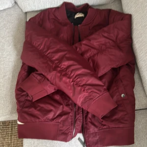 Vinröd bomberjacka från Crocker - Säljer en vinröd bomberjacka från Crocker i storlek L. Jackan har dragkedja framtill, ribbade muddar och svart quiltat innerfoder. Klassisk modell med fickor på sidorna och en stilren look.