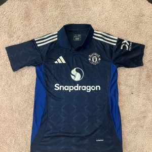 Manchester United bortatröja 2023/24 - Säljer en mörkblå Manchester United bortatröja från Adidas med guldiga detaljer och loggor. Tröjan har korta ärmar, klassisk krage och Snapdragons sponsortryck på bröstet. Slim fit-modell i lätt och ventilerande material.