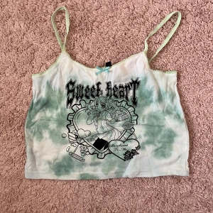 Grön och vit linnetopp  - Säljer en söt linnetopp från NEW girl ORDER i grönt och vitt batikmönster. Toppen har tunna axelband, liten rosett framtill och ett tryck med anime-inspirerad figur och texten 'Sweet heart'. Perfekt för en unik och lekfull stil! Köpt på ASOS, använd en gång. Skulle säga storlek M