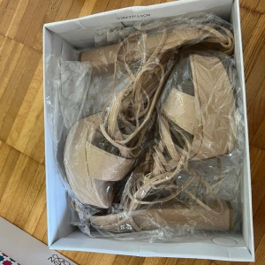 Beiga platåsandaler med snörning och klack - Säljer ett par snygga beiga sandaler med hög klack och platåsula. Skorna har öppen tå och smala remmar med snörning som går upp runt vristen. 