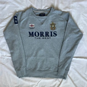 Grå sweatshirt från Morris  - Säljer en grå sweatshirt från Morris med broderad logga och text på bröstet. Tröjan har rund hals, långa ärmar och tryckt siffra på ena ärmen. Mjukt material och klassisk passform.
