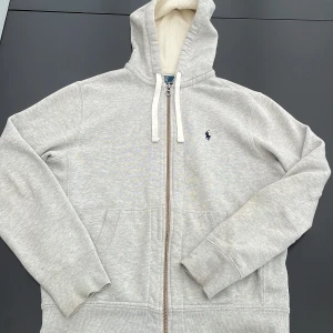 Grå hoodie från Polo Ralph Lauren - Säljer en ljusgrå hoodie från Polo Ralph Lauren med dragkedja och vit dragsko i huvan. Tröjan har broderad logga på bröstet och två fickor framtill. Perfekt för en avslappnad stil. Lite missfärgat på vänstra underarmen( bild två ser man det på)