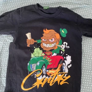 Svart t-shirt med tecknad tryck från Cortez - Cool svart t-shirt med färgglatt tecknat tryck av en figur i en grön och röd bil, eldflammor och Critiz-logga i gult. Perfekt för dig som gillar unika och uttrycksfulla plagg.