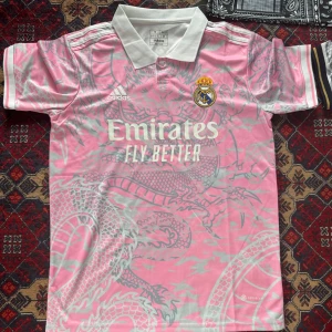 Real Madrid rosa fotbollströja med drakmönster - Säljer en rosa Real Madrid fotbollströja från Adidas med vitt drakmönster och klubbmärke på bröstet. Tröjan har vit krage och korta ärmar. Emirates Fly Better-logga på framsidan.Storleken på tröjan är S, De är helt nya passa på köp, du kan även beställa andra fotbollströjor av oss de ba o skriva privat till oss så löser vi, köparen står för frakten.